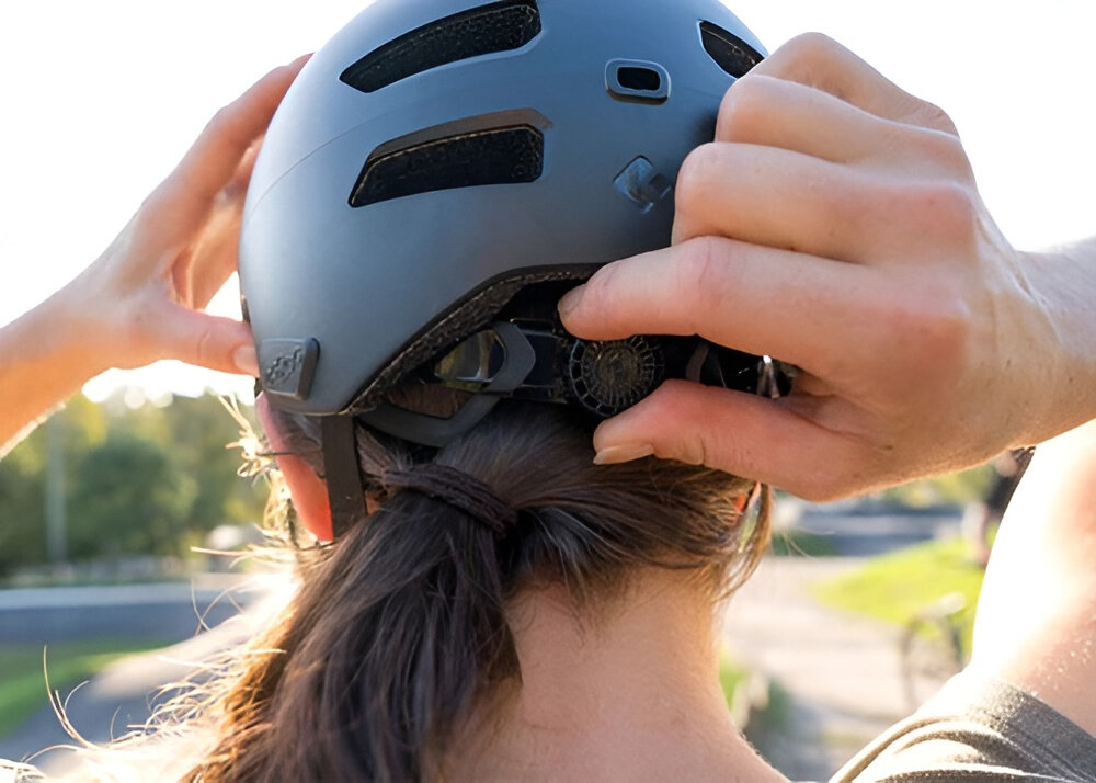Osoba reguluje pasek kasku rowerowego na głowie. Kask jest w kolorze białym matowym z widocznymi otworami wentylacyjnymi.