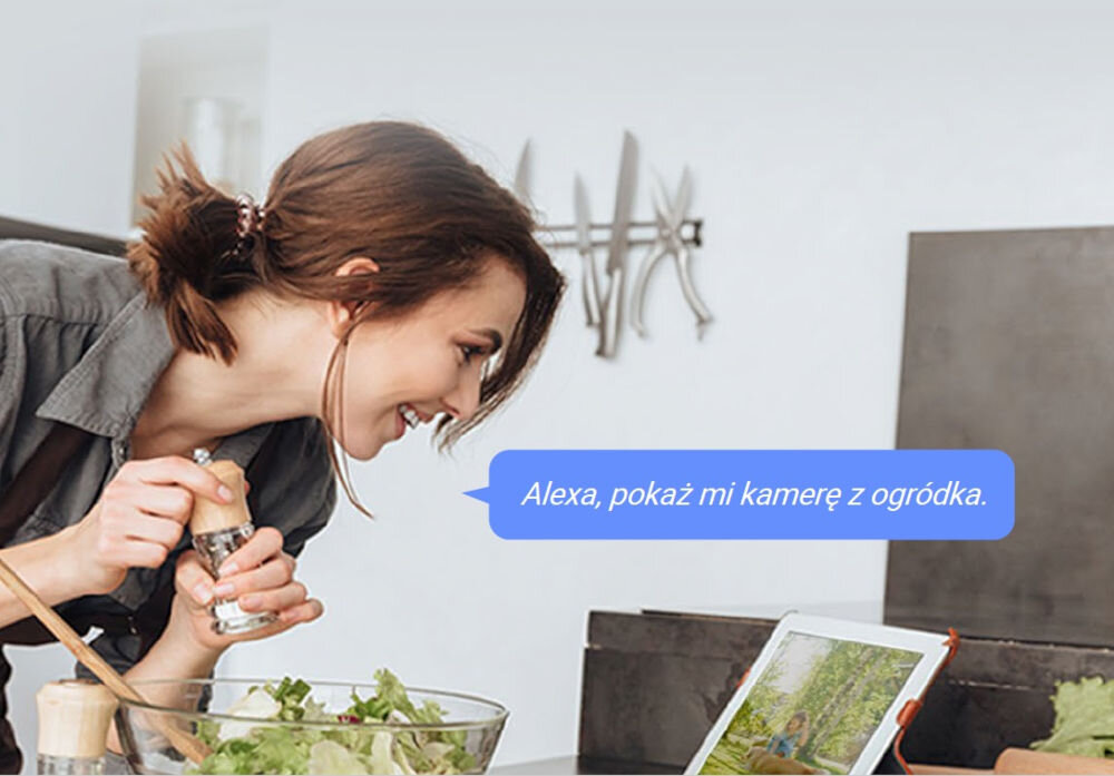 Kobieta w kuchni przyprawia sałatkę, patrząc na tablet, na którym wyświetlany jest obraz z kamery. Na ekranie widoczny jest tekst: \'Alexa, pokaż mi kamerę z ogródka.\'