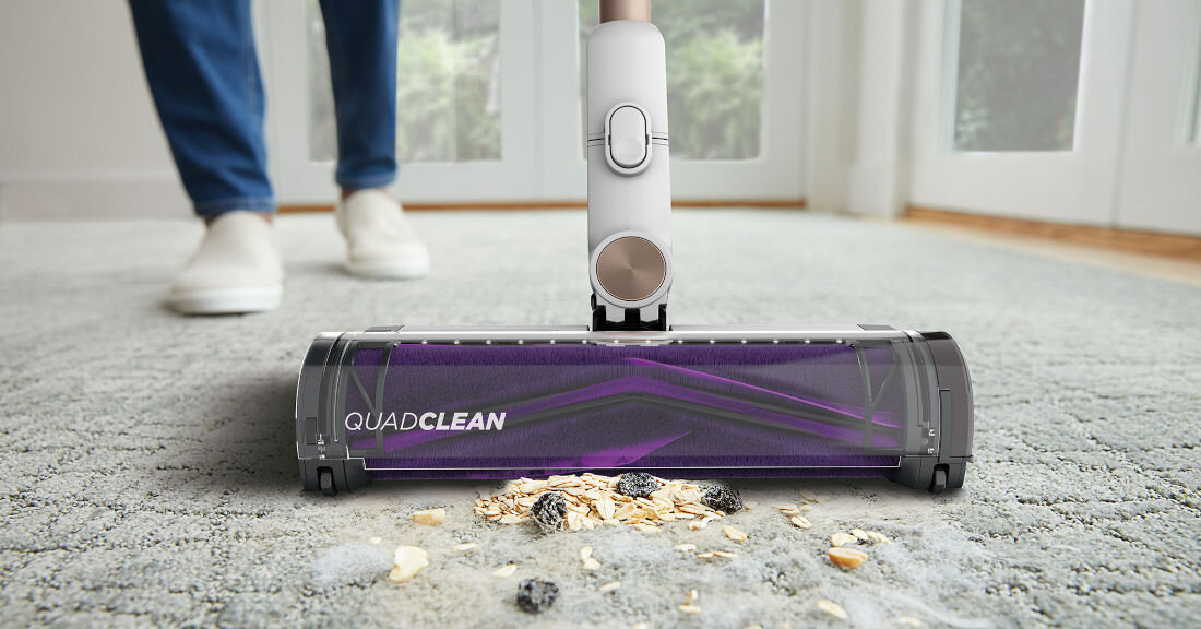 Odkurzacz z napisem \'QUADCLEAN\' na głowicy, zbiera zanieczyszczenia z dywanu. W tle widoczne są drzwi balkonowe i osoba w białych butach.