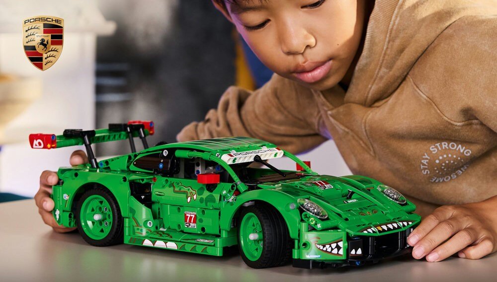 Na obrazie znajduje się zielony model samochodu z klocków LEGO, z widocznymi detalami i naklejkami, obok którego znajduje się dziecko. W tle widoczne jest logo Porsche.