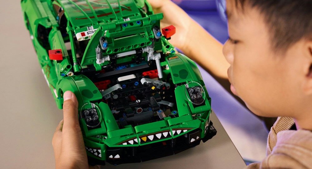Osoba trzyma zielony model samochodu z klocków LEGO z otwartą maską, ukazującą szczegóły konstrukcji. Na masce widoczne są naklejki z wzorem przypominającym zęby.