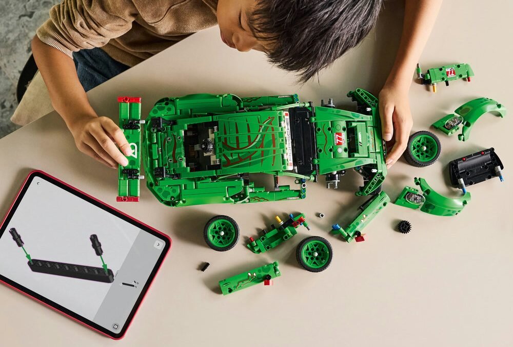 Osoba składa zielony model samochodu z klocków LEGO, wokół którego leżą różne części. Obok znajduje się tablet z instrukcją montażu wyświetloną na ekranie.
