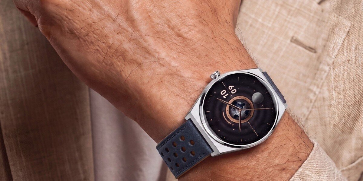 Osoba nosi smartwatch z granatowym paskiem i okrągłą tarczą, na której widoczne są cyfry i wskazówki. Tło stanowi jasna marynarka.