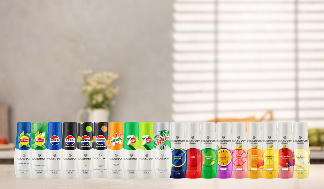 Na obrazie widocznych jest szesnaście butelek syropów do saturatorów marki SodaStream, w różnych kolorach i smakach, ustawionych w rzędzie. W tle znajduje się rozmyty widok kuchni z rośliną i oknem z żaluzjami.