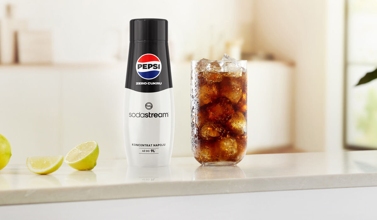Na blacie stoi butelka z napisem \'Pepsi Zero Cukru sodastream\' oraz szklanka z napojem i kostkami lodu. Obok leżą kawałki limonki.