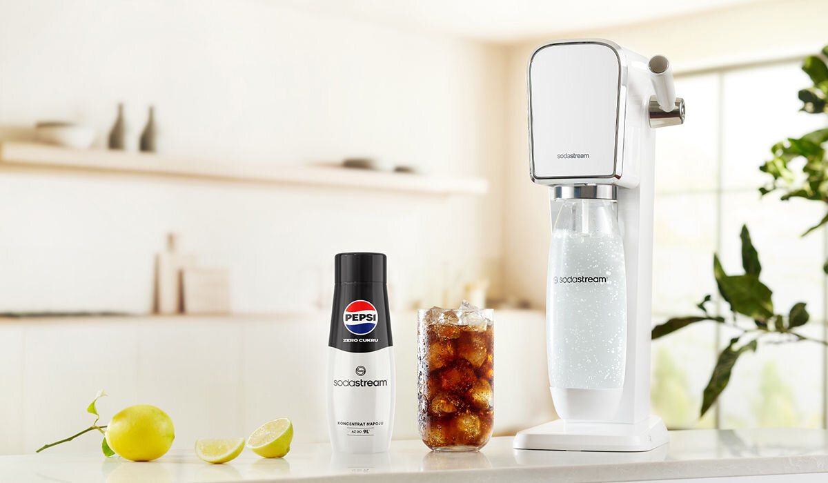 Na blacie kuchennym znajduje się biały saturator marki sodastream z butelką wypełnioną gazowaną wodą. Obok stoi butelka syropu Pepsi Zero Cukru oraz szklanka z napojem i kostkami lodu.