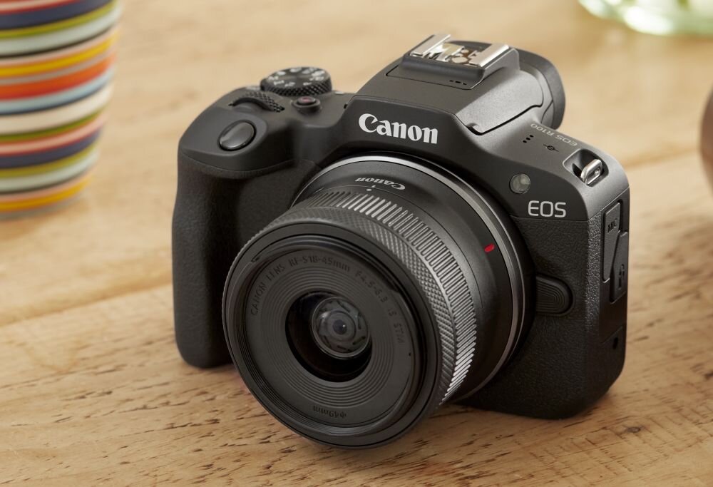 Na drewnianym stole leży aparat bezlusterkowy z logo Canon i oznaczeniem EOS. Obiektyw ma widoczny napis \'Canon Lens RF-S 18-45mm\'.