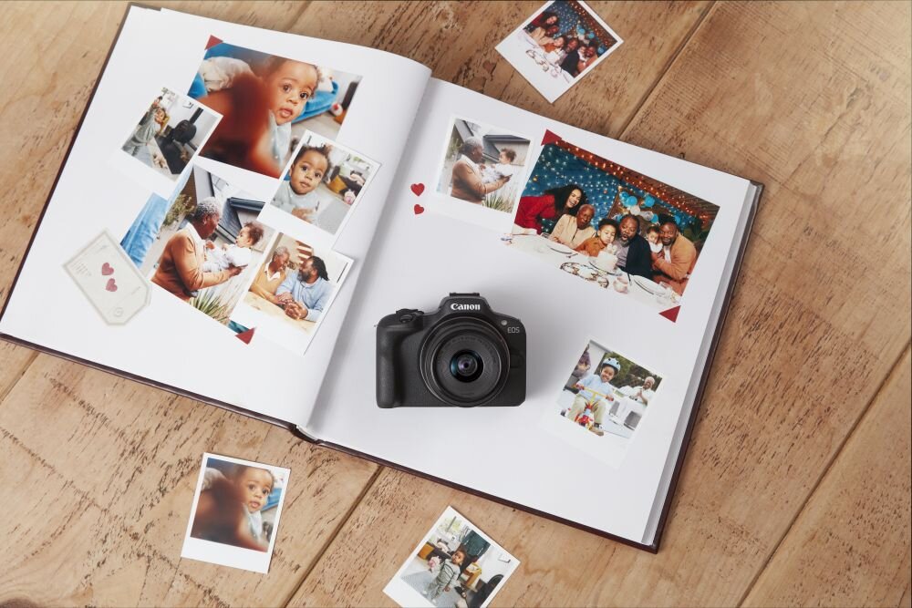 Na drewnianym stole leży otwarty album ze zdjęciami, wokół którego rozłożone są dodatkowe fotografie. Na albumie znajduje się aparat z widocznym logo \'Canon\'.
