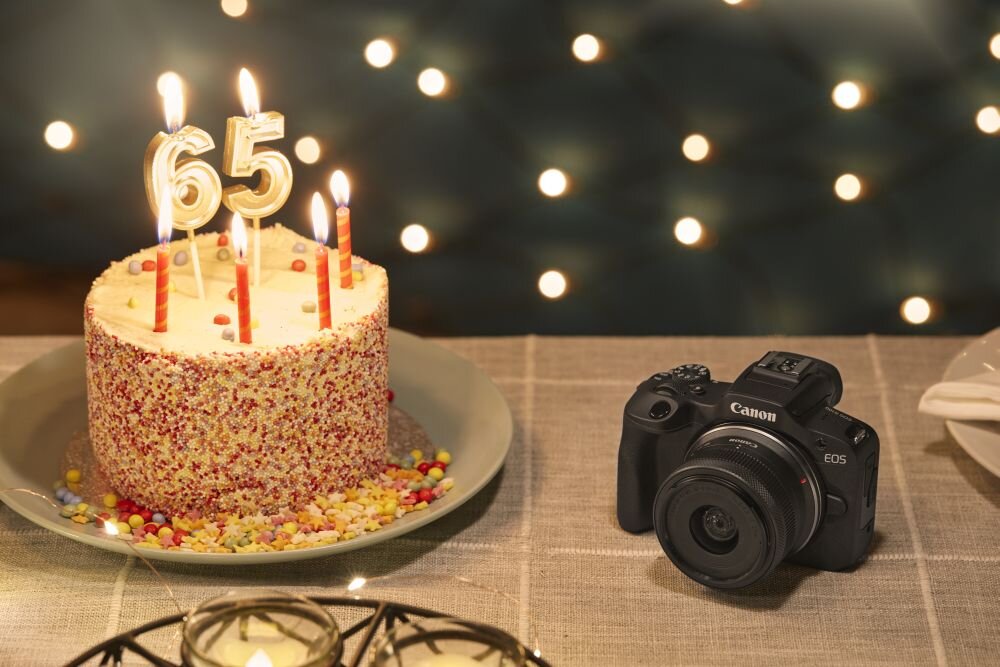 Na stole znajduje się tort z zapalonymi świeczkami w kształcie liczby 65 oraz aparat fotograficzny z logo Canon EOS. W tle widoczne są rozmyte światła, tworzące efekt bokeh.