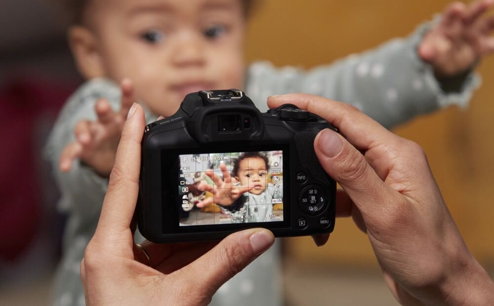 Osoba trzyma aparat fotograficzny, na ekranie którego widać dziecko wyciągające ręce w stronę obiektywu. W tle znajduje się rozmyta postać dziecka w podobnej pozycji.