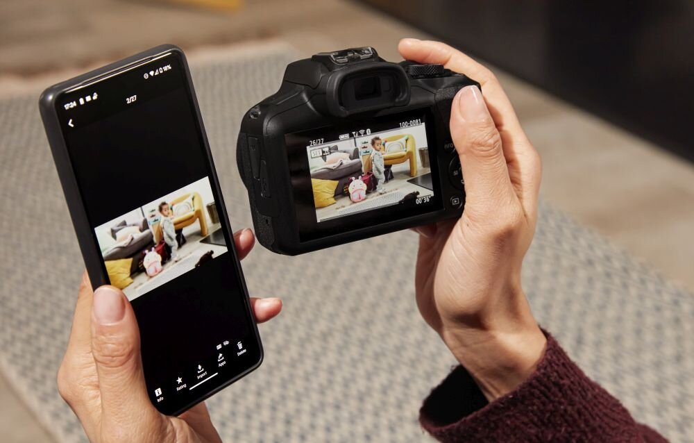Osoba trzyma aparat fotograficzny i smartfon, na obu ekranach widoczny jest ten sam obraz przedstawiający dziecko siedzące na kanapie z plecakiem. Aparat ma duży wyświetlacz, a smartfon pokazuje zdjęcie w aplikacji galerii.