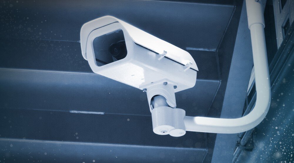Obraz przedstawia zamontowaną kamerę monitoringu w białej obudowie, skierowaną w dół. Kamera znajduje się na tle metalowej konstrukcji.