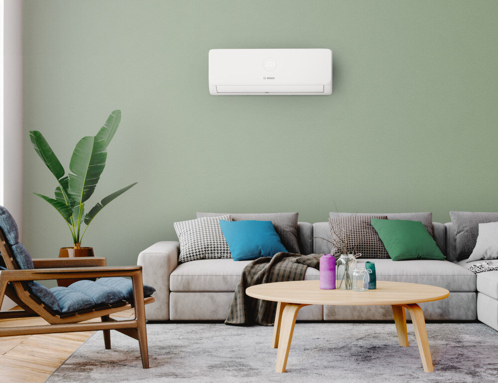 W salonie na ścianie zamontowany jest biały klimatyzator z logo \'BOSCH\'. Przed nim znajduje się szara sofa z kolorowymi poduszkami, drewniany stolik oraz roślina w doniczce.