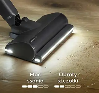 Na drewnianej podłodze znajduje się końcówka odkurzacza z podświetleniem, zbierająca rozsypane drobinki. Widoczny jest tekst: \'Moc ssania\' z trzema białymi prostokątami oraz \'Obroty szczotki\' z dwoma białymi prostokątami.