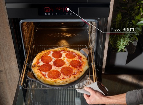 Otwarty piekarnik z pizzą na ruszcie, obok widoczna ręka trzymająca uchwyt. Na wyświetlaczu piekarnika widoczny jest napis \'Pizza 300°C\'.