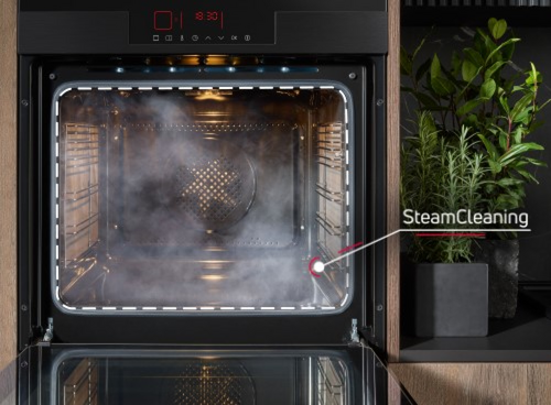 Otwarty piekarnik z funkcją SteamCleaning, z wnętrza wydobywa się para. Obok piekarnika znajduje się doniczka z zieloną rośliną.