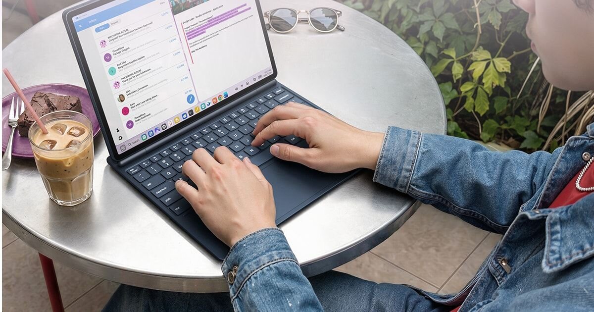 Osoba używa tabletu z klawiaturą na okrągłym stole, obok stoi szklanka z kawą. Na ekranie widoczne są aplikacje do komunikacji i edycji tekstu.