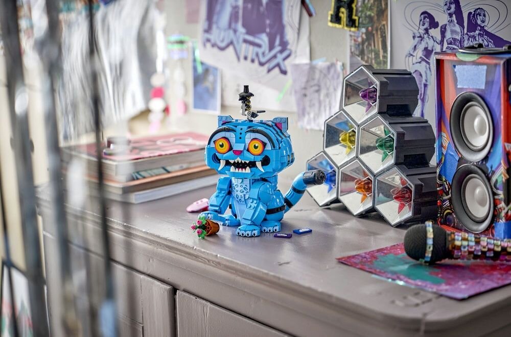 Na biurku znajduje się niebieska figurka LEGO przedstawiająca tygrysa z dużymi oczami i otwartą paszczą. Obok niej leży mikrofon, a w tle widać głośniki i kolorowe plakaty na ścianie.