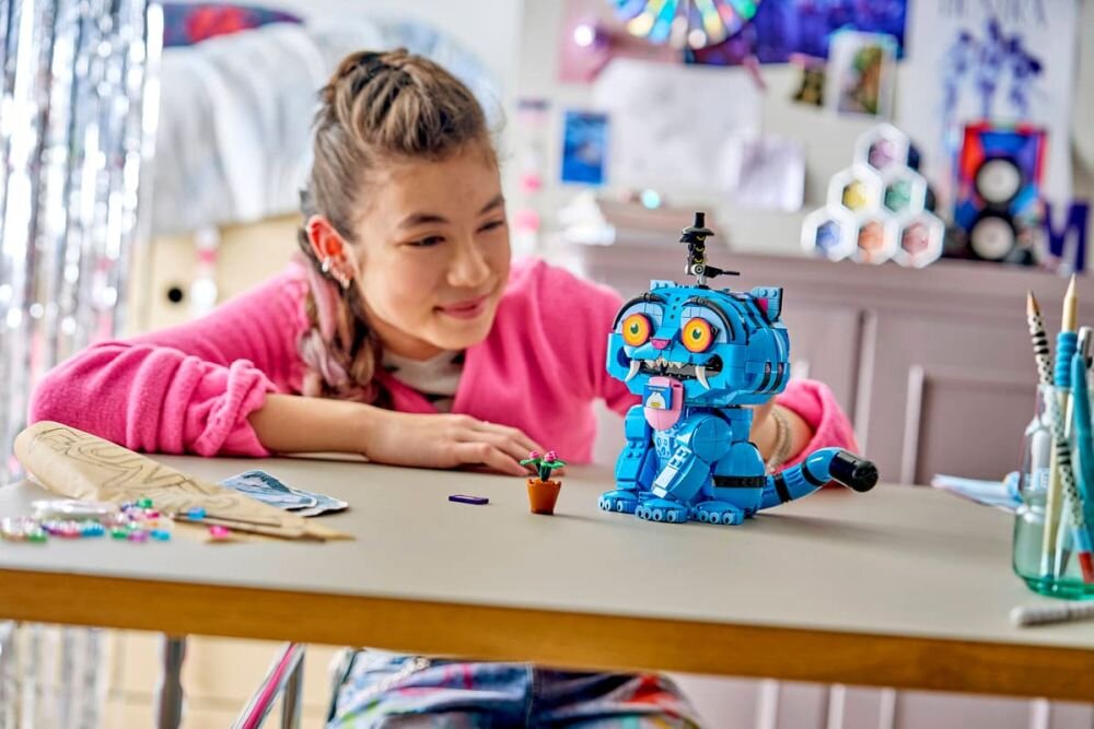 Osoba w różowej bluzie siedzi przy stole, na którym znajduje się niebieski model LEGO przypominający kota z dużymi oczami i otwartą paszczą. W tle widać rozmyte elementy dekoracyjne i meble.