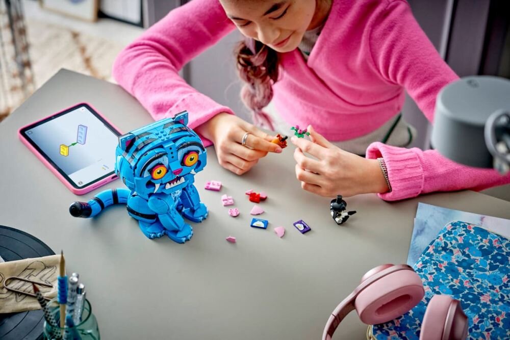 Osoba w różowym swetrze bawi się zestawem klocków LEGO, w tym niebieskim modelem tygrysa. Na stole leży tablet z instrukcją oraz różowe słuchawki.