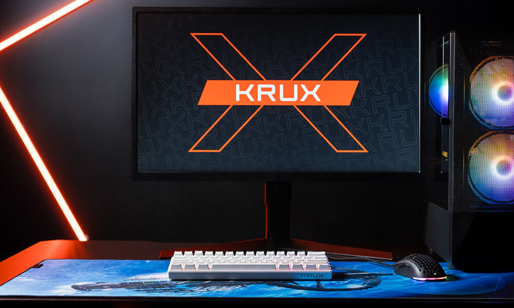 Na biurku znajduje się biała klawiatura oraz myszka na podkładce z niebieskim wzorem. W tle widoczny jest monitor z logo \'KRUX\' oraz komputer z podświetleniem RGB.
