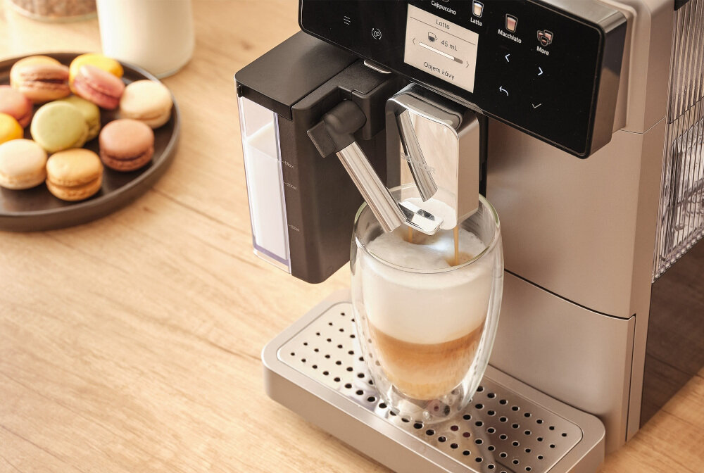 Ekspres do kawy przygotowuje napój w szklance z pianką, a obok na stole znajduje się talerz z kolorowymi makaronikami. Na panelu ekspresu widoczny jest tekst: \'Cappuccino\', \'Latte\', \'Macchiato\', \'More\', \'Dławn 60s\'.