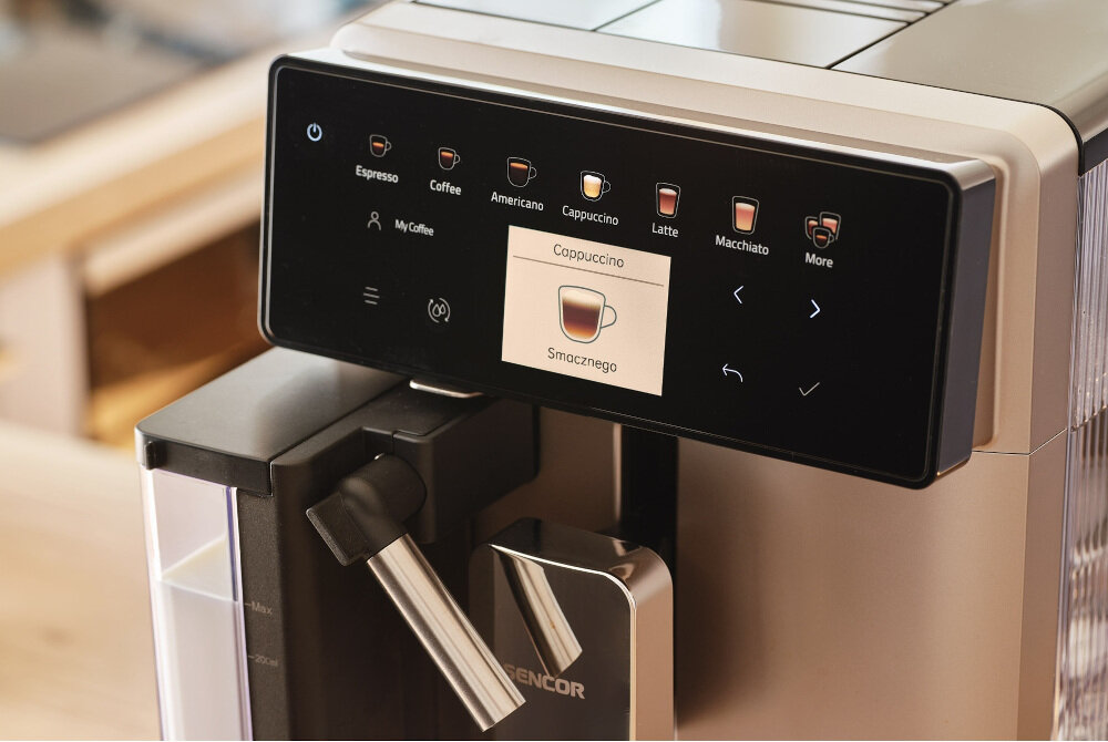 Ekspres do kawy z panelem dotykowym, na którym widoczne są opcje takie jak Espresso, Coffee, Americano, Cappuccino, Latte, Macchiato oraz przycisk \'More\'. Na ekranie wyświetla się napis \'Cappuccino Smacznego\'.