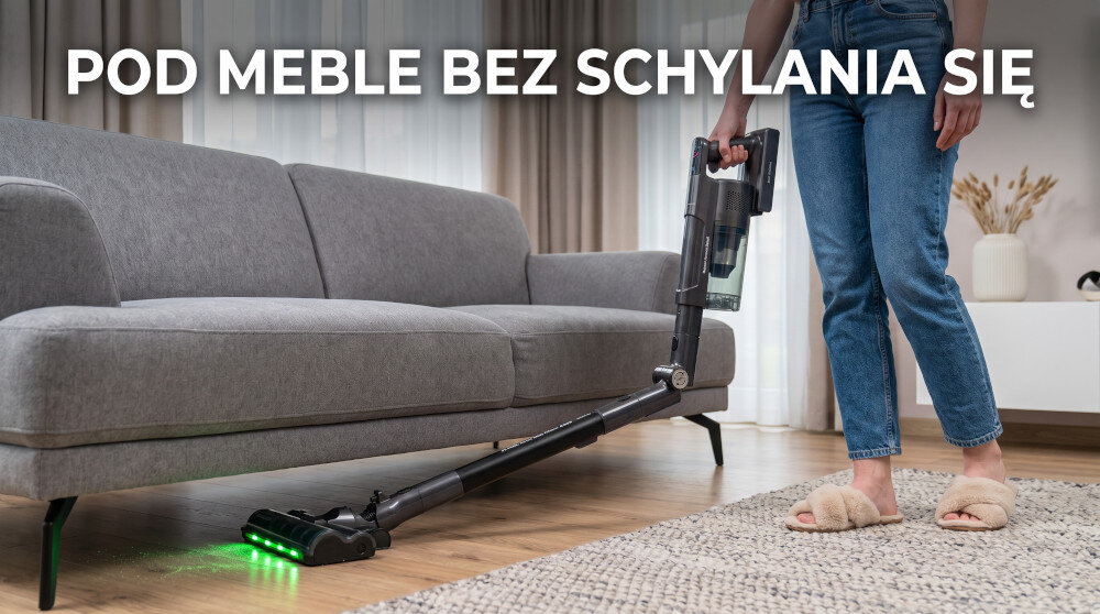Osoba używa odkurzacza pionowego z elastyczną rurą, który wsuwa się pod szarą sofę. Na obrazie widoczny jest tekst: \'POD MEBLE BEZ SCHYLANIA SIĘ\'.