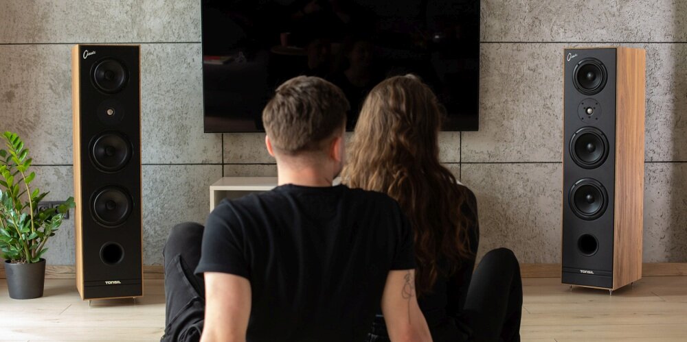 Na obrazie widoczne są dwie kolumny głośnikowe Tonsil Maestro Classic w kolorze czarnym, ustawione w pomieszczeniu z telewizorem na ścianie. Przed nimi siedzą dwie osoby, a obok jednej z kolumn stoi roślina w doniczce.