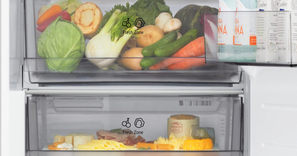Wnętrze lodówki z dwoma szufladami oznaczonymi jako \'Fresh Zone\', wypełnionymi warzywami i produktami spożywczymi. Na drzwiach widoczne są kartony z napojami.