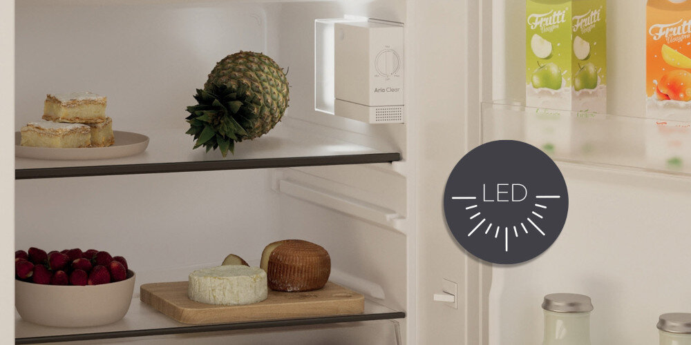Wnętrze lodówki z widocznymi półkami, na których znajdują się ananas, ser, ciasto i miska z owocami. Na drzwiach lodówki widoczne są kartony z sokami, a obok znajduje się ikona z napisem \'LED\'.