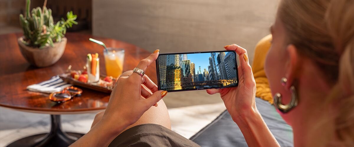 Osoba trzyma smartfon, na którym wyświetlany jest obraz miasta z wieżowcami. W tle widać stolik z napojem i kaktusem.