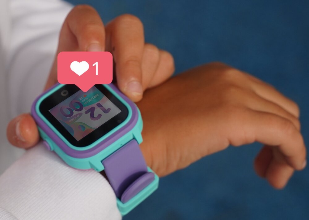 Osoba nosi smartwatch z niebiesko-fioletowym paskiem, na ekranie widoczna jest godzina 12:00. Nad zegarkiem unosi się ikona serca z liczbą 1.