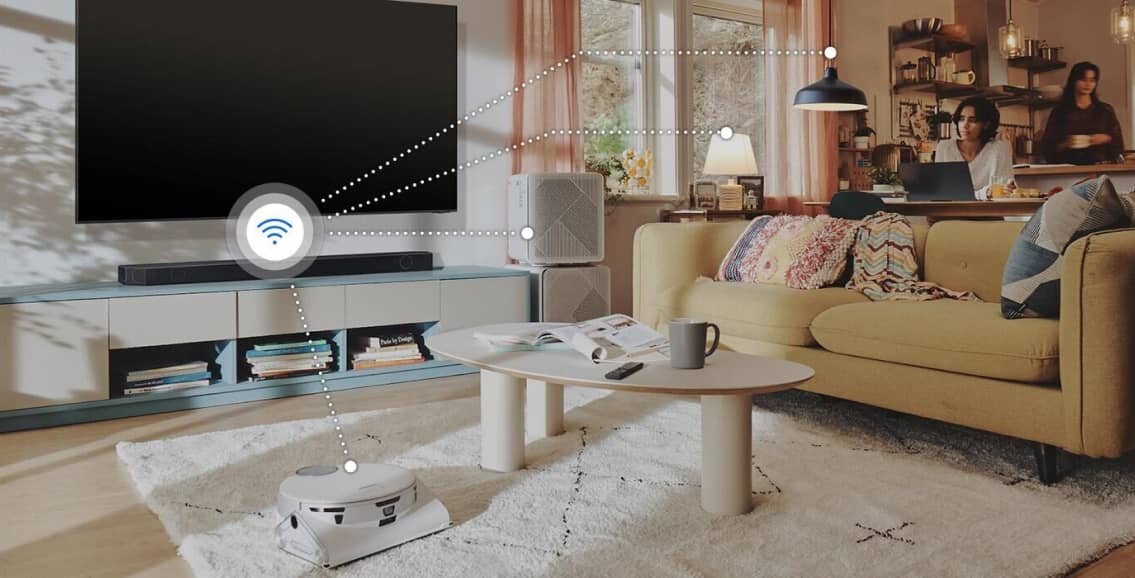 W salonie znajduje się duży telewizor na szafce, przed którym stoi soundbar. Na dywanie leży robot sprzątający, a w tle widać dwie osoby przy stole w kuchni.