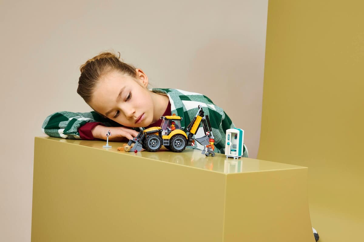 Na obrazie widać dziecko pochylone nad żółtą koparko-ładowarką LEGO oraz kilkoma figurkami i akcesoriami. Tło jest jednolite, w kolorze beżowym.