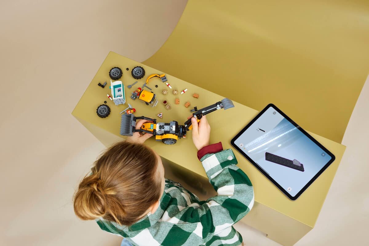 Osoba składa model LEGO żółtej koparko-ładowarki na jasnym stole, obok leży tablet z wyświetlonym obrazem klocka LEGO. Na stole znajdują się również inne elementy zestawu, takie jak koła i małe figurki.