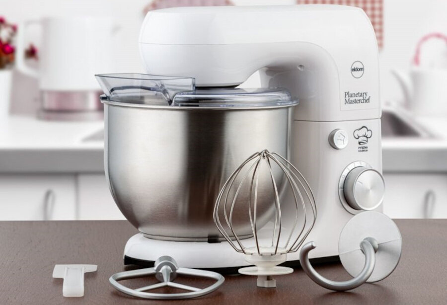 Na obrazie znajduje się biały robot kuchenny z metalową misą i trzema różnymi końcówkami do mieszania. Na urządzeniu widoczne jest logo \'eldom\' oraz napis \'Planetary Masterchef\'.
