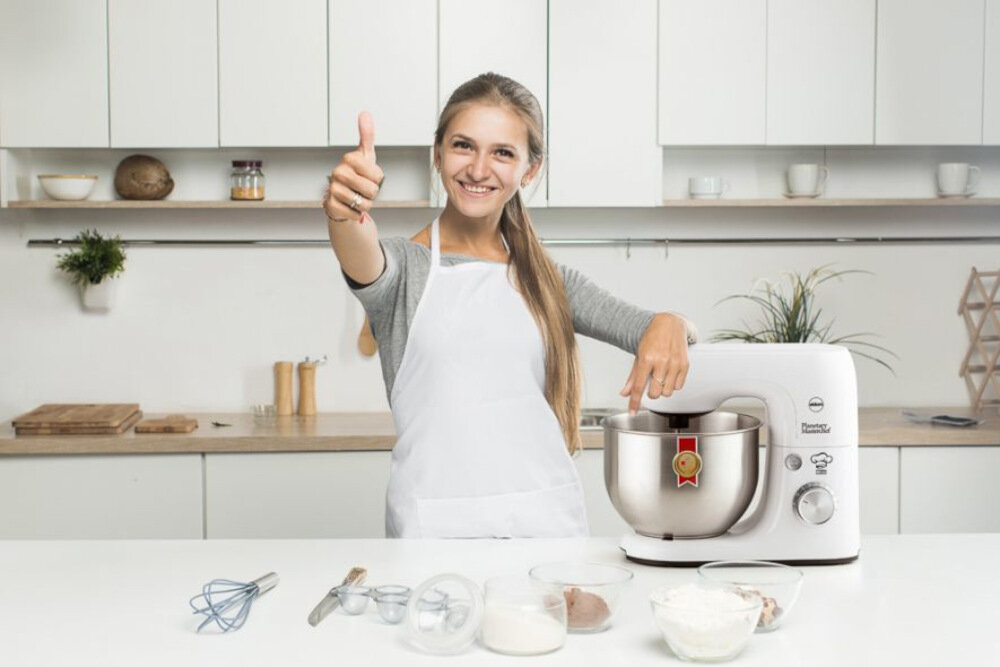 Kobieta w fartuchu stoi obok robota kuchennego, pokazując gest kciuka w górę. Na blacie znajdują się różne akcesoria kuchenne i składniki.