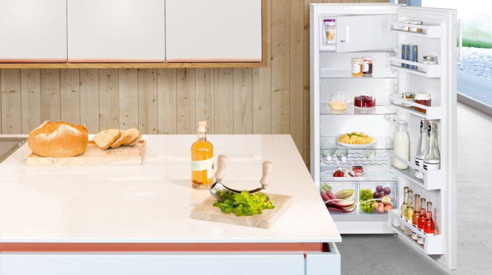 Kuchnia z otwartą białą lodówką wypełnioną różnymi produktami spożywczymi, obok której znajduje się blat z chlebem, butelką napoju i deską z zielonymi liśćmi.