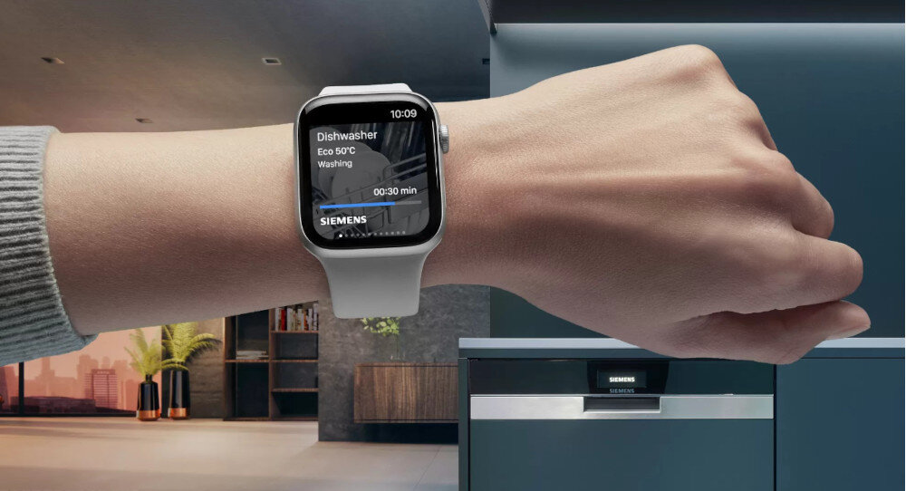 Osoba nosi smartwatch z wyświetlaczem pokazującym informacje o zmywarce: \'Dishwasher Eco 50°C Washing 00:30 min\' oraz logo \'SIEMENS\'. W tle widoczna jest zmywarka z panelem sterowania i logo \'SIEMENS\'.