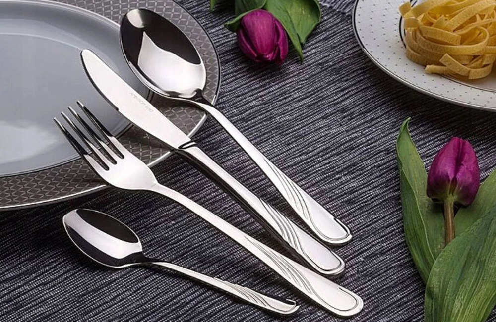 Na obrazie widoczny jest zestaw sztućców składający się z noża, widelca, łyżki i łyżeczki, ułożonych na ciemnym tle obok talerza z makaronem i tulipanów. Sztućce mają elegancki, metaliczny połysk.