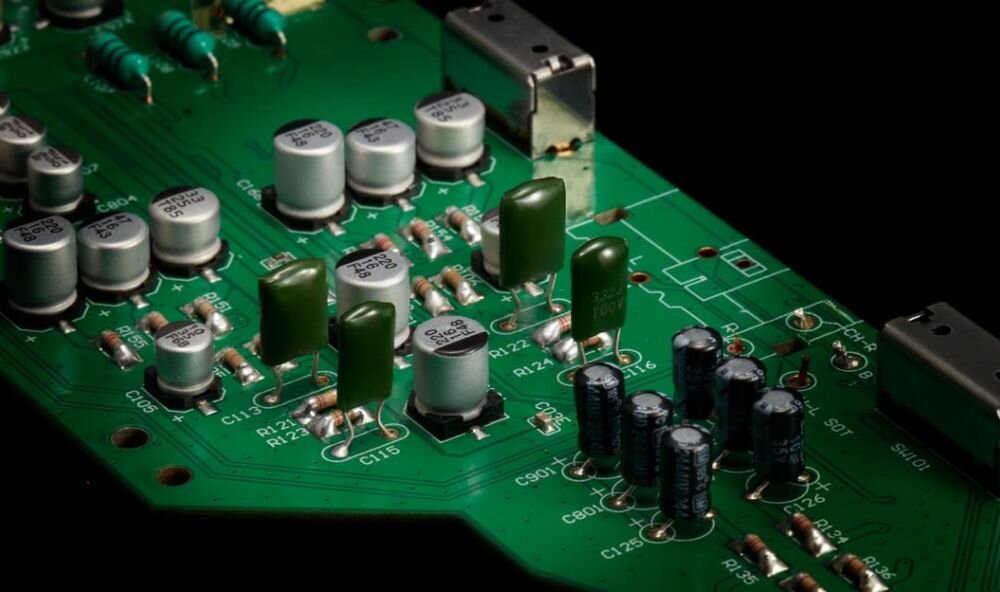 Zdjęcie przedstawia zieloną płytkę drukowaną z różnymi elementami elektronicznymi, takimi jak kondensatory i rezystory. Widoczne są także metalowe złącza na krawędziach płytki.