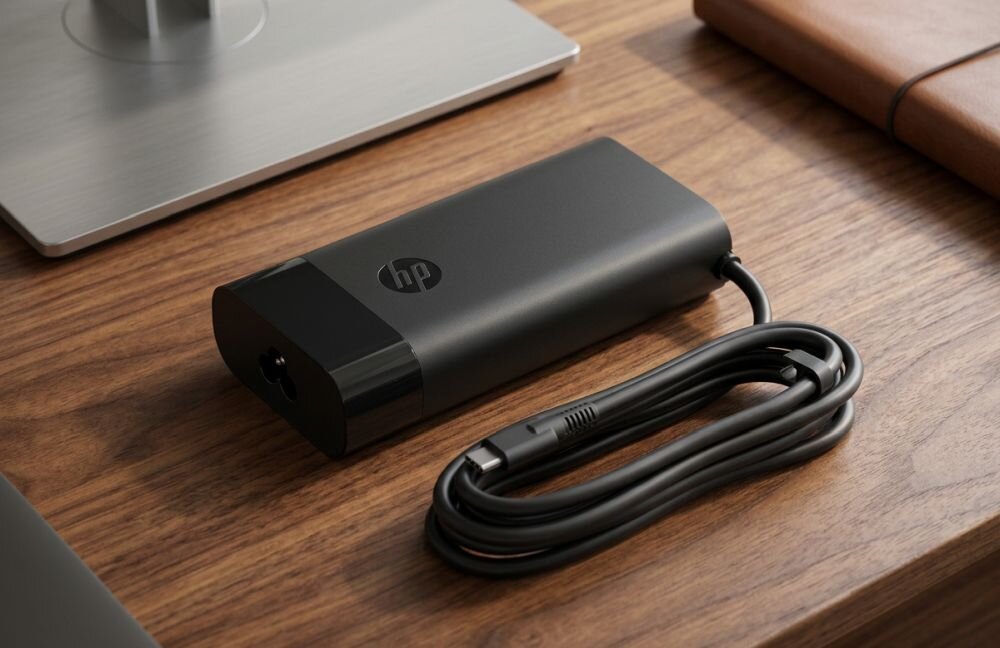 Na drewnianym blacie znajduje się czarny zasilacz z logo HP oraz zwinięty kabel USB-C.