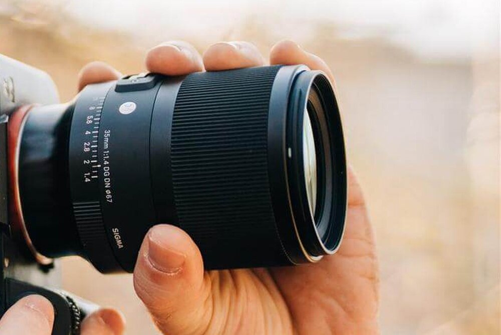 Osoba trzyma obiektyw fotograficzny z widocznymi napisami: \'35mm f/1.4 DG DN Ø67\' oraz \'SIGMA\'.