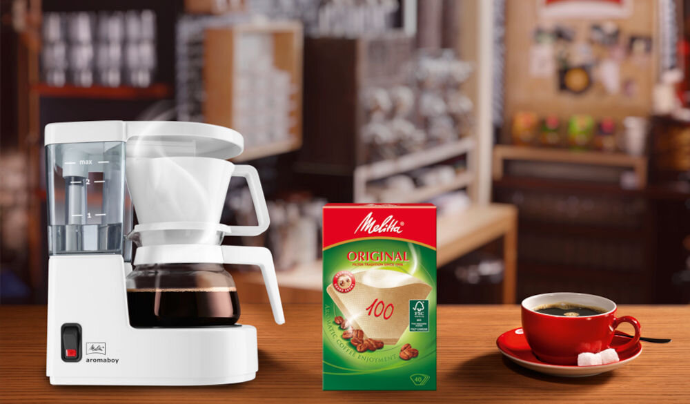 Na blacie stoi biały ekspres do kawy z napisem \'aromaboy\', obok znajduje się opakowanie filtrów do kawy z logo \'Melitta\'. W tle widoczna jest rozmyta kawiarnia, a na blacie stoi czerwona filiżanka z kawą.