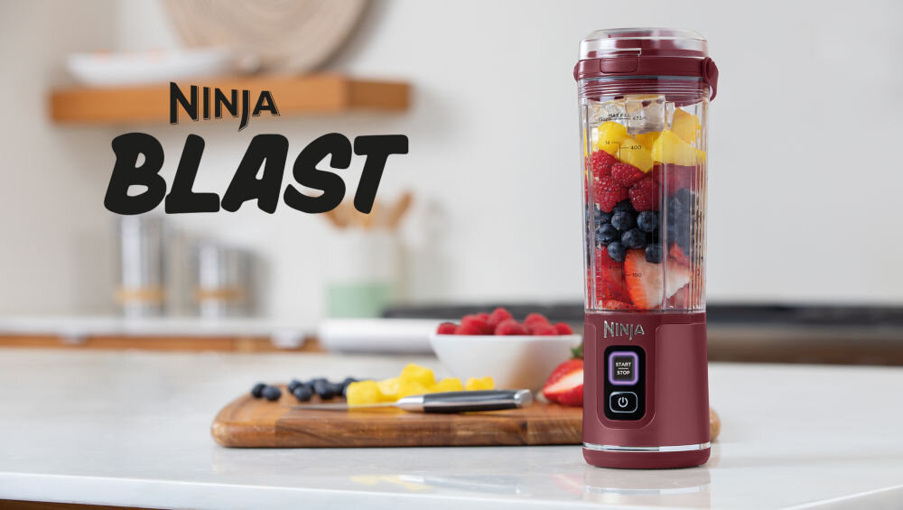 Na blacie kuchennym stoi czerwony blender z logo \'NINJA\', wypełniony owocami. Obok znajduje się deska z pokrojonymi owocami i nożem, a w tle widoczny jest napis \'BLAST\'.