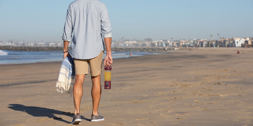 Osoba idzie po plaży, trzymając w ręku czerwony blender personalny z napojem. W tle widać zabudowania i morze.