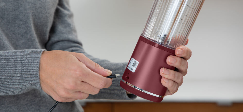 Osoba trzyma czerwony blender personalny i podłącza do niego kabel. Blender ma przezroczysty pojemnik z miarką.