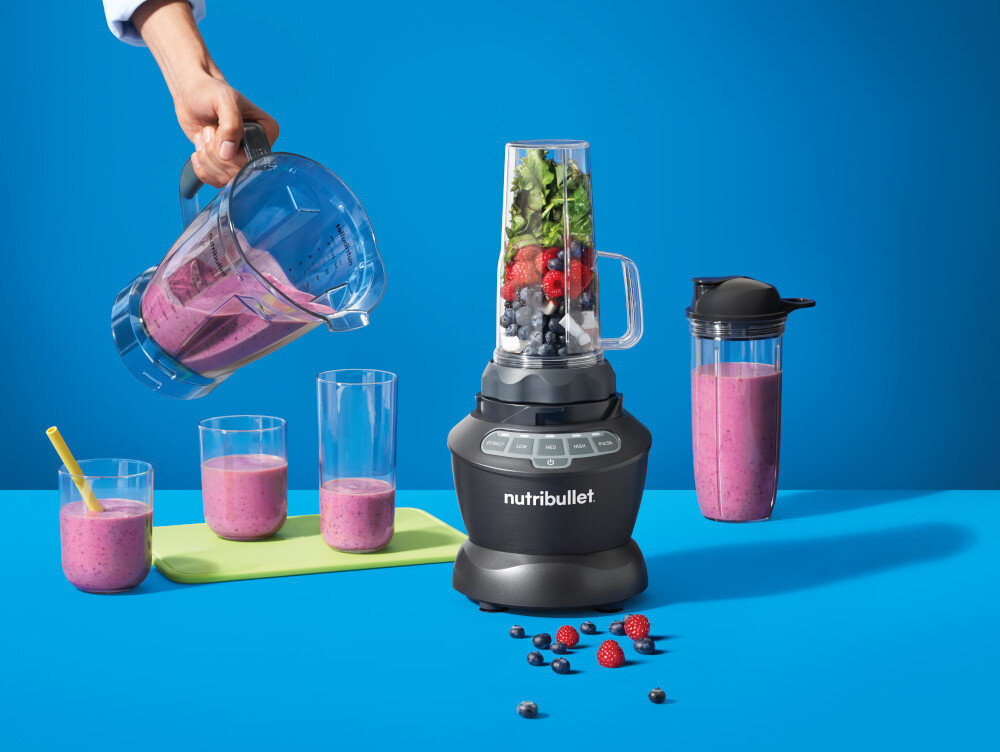 Na niebieskim tle znajduje się blender kielichowy z logo \'nutribullet\', wypełniony owocami i warzywami. Obok stoją szklanki z różowym napojem oraz dzbanek, z którego ktoś nalewa płyn.