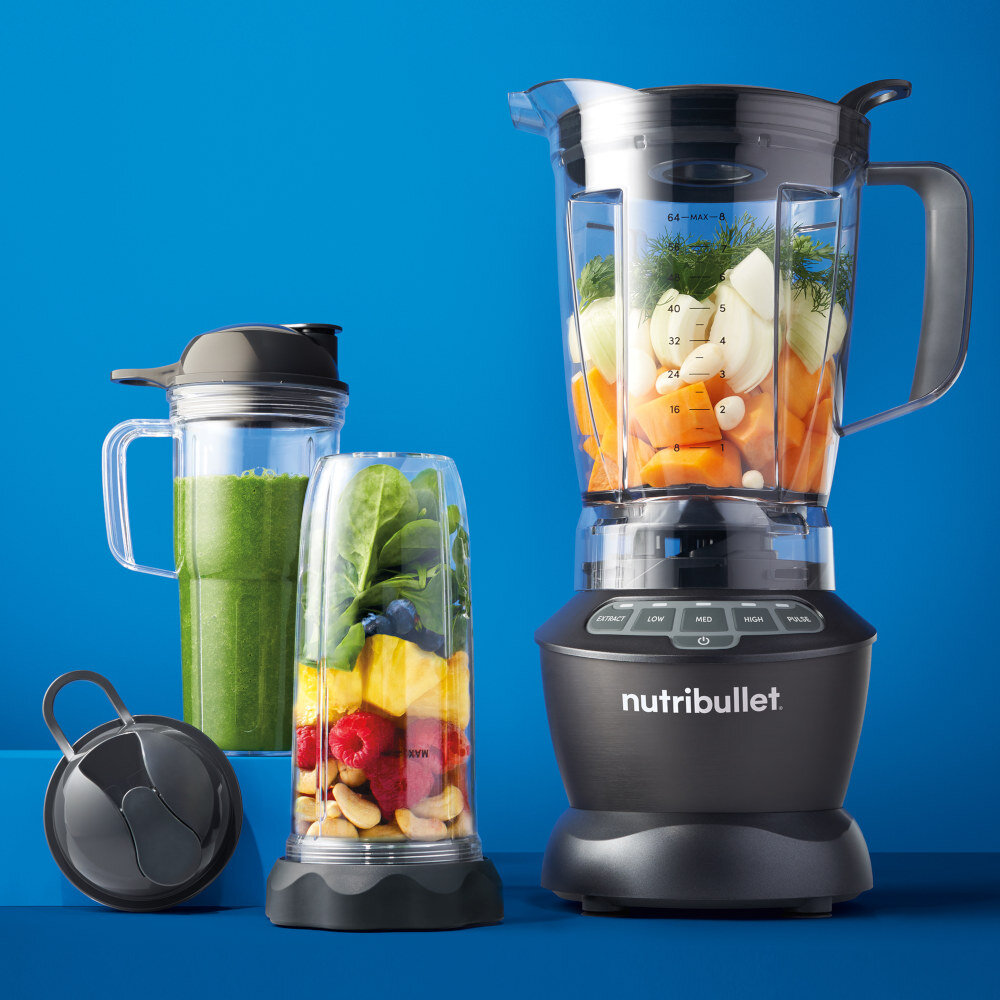 Na niebieskim tle znajduje się blender kielichowy z logo \'nutribullet\', wypełniony warzywami. Obok stoją dwa mniejsze pojemniki z pokrywkami, jeden z zielonym napojem, drugi z owocami i orzechami.